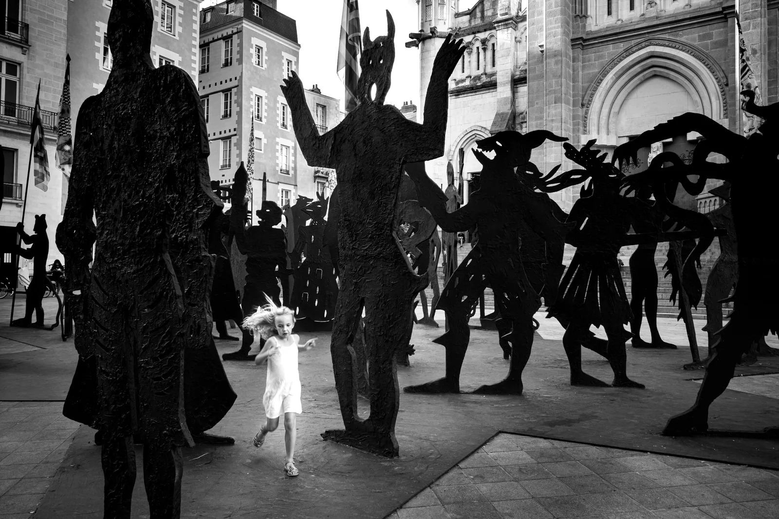 A girl runs through a crowd of dark iron sculptures in Nantes — a photograph from the Nantais et Al exhibition by Dan Umareta